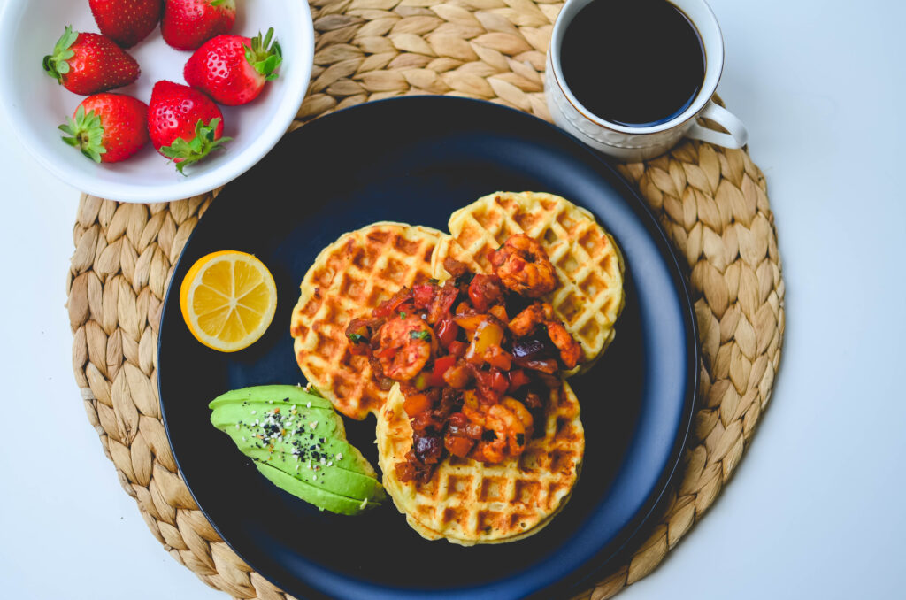 A overhead shot of cassava corn waffles with avocado, shrimp creole with fruit and coffee